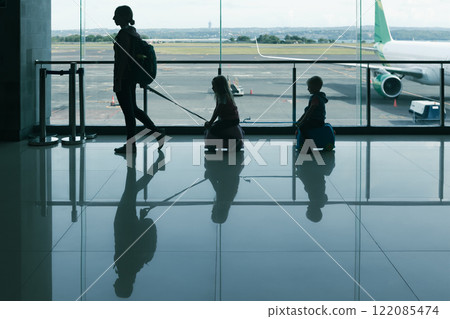 A mother pulls two children on small ride-on luggage through airport terminal, silhouetted against large windows with airplane in view. This scene captures family travel and fun moments before A mother pulls two children on small ride-on luggage through airport terminal, silhouetted against large windows with airplane in view. This scene captures family travel and fun moments before 122085474