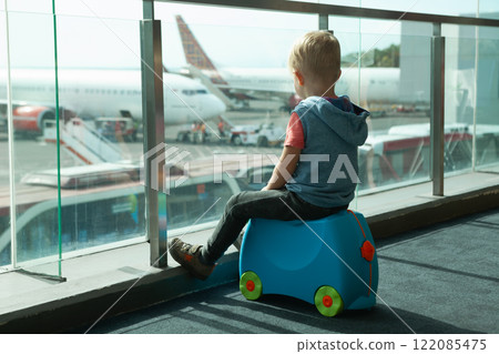 Young boy sitting on a ride-on suitcase, looking out of the airport window at airplanes on the runway. Capturing the anticipation and excitement of family travel and exploration. 122085475
