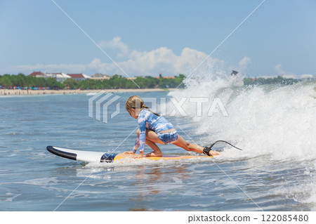 Happy baby girl - young surfer ride on surfboard with fun on sea waves. Active family lifestyle, kids outdoor water sport lessons and swimming activity in surf camp. Summer vacation with child. 122085480