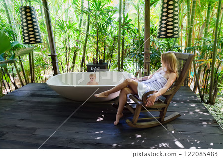 Happy baby boy have fun in bathtub, young mother relax on rocking chair in outside bathroom on veranda with beautiful tropical garden view at luxury villa on summer family vacation. 122085483