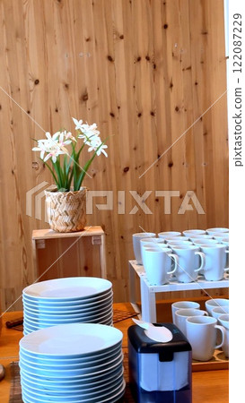 Warm wooden tables adorned with vibrant flower in eco-friendly cafe.Clean plates and cups are thoughtfully arranged, inviting guests to enjoy nature-inspired dining experience Eco-friendly ambiance  122087229