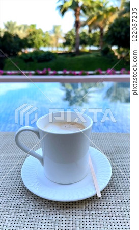 Coffee cup on reflective glass table, coffee blending with reflections of palm trees outdoor greenery, creating serene outdoor ambiance where reflections and calm moments invite peaceful relaxation  122087235