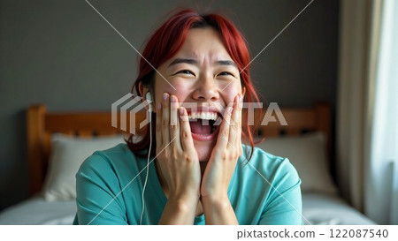 An excited, laughing, positive Asian woman wearing headphones, on a bed, close-up, making an audio call 122087540