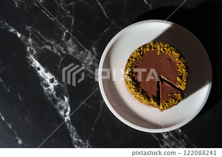 Top view of Dubai chocolate cheesecake with pistachio paste and kataifi covered in chocolate ganache on white plate. Black background with copy space. Indulgence, gourmet desserts, art of culinary Top view of Dubai chocolate cheesecake with pistachio paste and kataifi covered in chocolate ganache on white plate. Black background with copy space. Indulgence, gourmet desserts, art of culinary 122088241