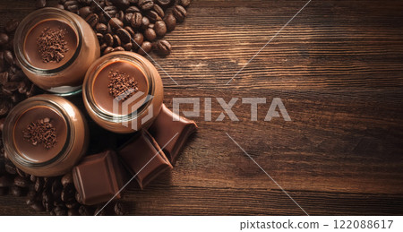 Mocha mousses in glass jars topped with chocolate on an old wooden table texture with fresh roasted coffee beans. Mix of brown shades as trend color for 2025 year 122088617