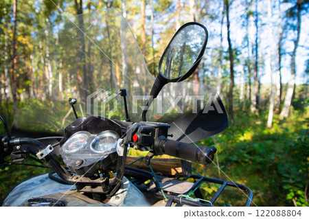 ATV windshield, steering wheel and rear view mirrors. Quad bike or ATV stands idle among tall trees and lush greenery. All Terrain Vehicle outside ATV windshield, steering wheel and rear view mirrors. Quad bike or ATV stands idle among tall trees and lush greenery. All Terrain Vehicle outside 122088804