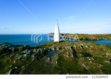 Seascape with structure. A tall, white, pointed tower is situated on a grassy, rocky headland. The ocean stretches to the horizon under a bright blue sky. Seascape with structure. A tall, white, pointed tower is situated on a grassy, rocky headland. The ocean stretches to the horizon under a bright blue sky. 122089405