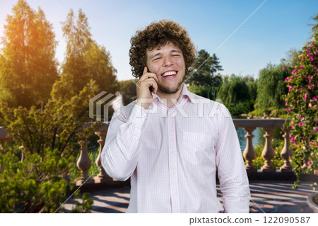 Portrait of a young man in white shirt talking on the phone and laughing. Green summer park in the background. 122090587