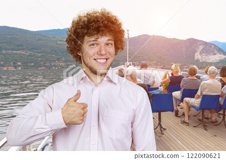Portrait of a happy young man with curly hair shows thumb up standing on the yacht board. Advertising a trip excursion. 122090621