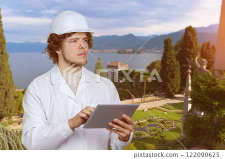 Portrait of a young man in white uniform and white helmet holding tablet pc device outdoors. Beautiful nature landscape in the background. 122090625