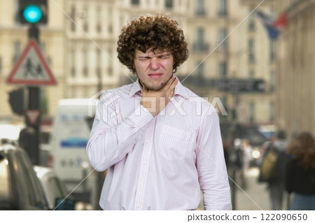Young handsome man with curly hair touching his neck. City street in the background. Young handsome man with curly hair touching his neck. City street in the background. 122090650