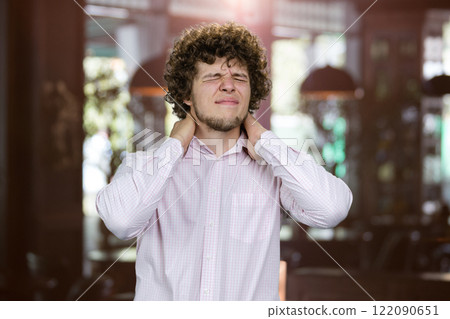 Young man with curly hair having neckache touching neck with both hands. Restaurant interior in the background. Young man with curly hair having neckache touching neck with both hands. Restaurant interior in the background. 122090651