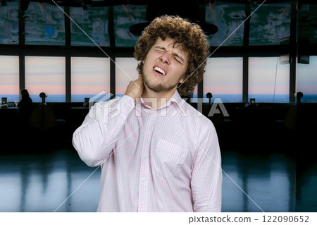 Young handsome man with curly hair having neckache. Dark indoor background. Young handsome man with curly hair having neckache. Dark indoor background. 122090652