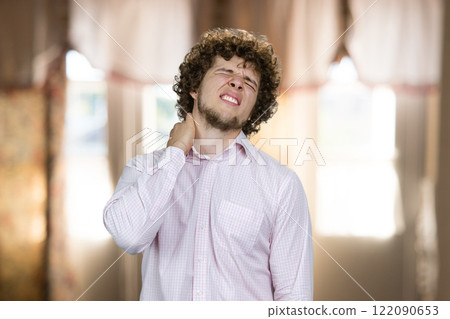 Young man with curly hair having neckache indoors. Home window curtains in the background. Young man with curly hair having neckache indoors. Home window curtains in the background. 122090653