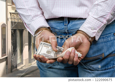 Close up of male hands in handcuffs holding dollar banknotes. Corruption and bribery concept. City street in the background. 122090675