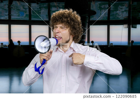 Portrait of an expressive young man with curly hair in white shirt screams in megaphone. 122090685