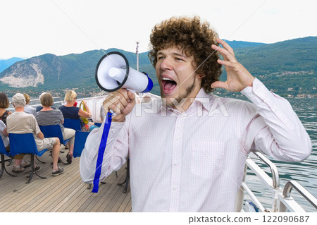 Portrait of a young excited expressive man with curly hair in white shirt screams out something in megaphone. Tourist people on the yacht boat in the background. 122090687