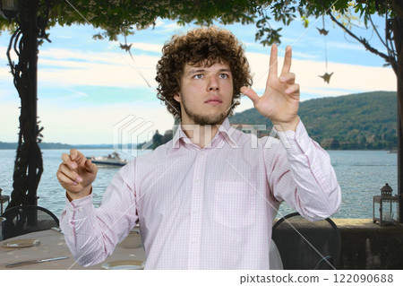 Portrait of a young man with curly hair and white shirt using invisible virtual screen for work outdoors. Sea resort beach in the background. 122090688