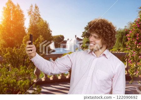 Portrait of happy young man with curly hair taking selfie with smartphone. City park in the background. Portrait of happy young man with curly hair taking selfie with smartphone. City park in the background. 122090692