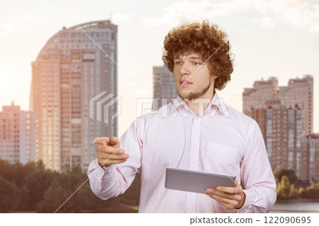 Young businessman with curly hair holding a tablet pc and pointing at something. Residential area in the background. 122090695