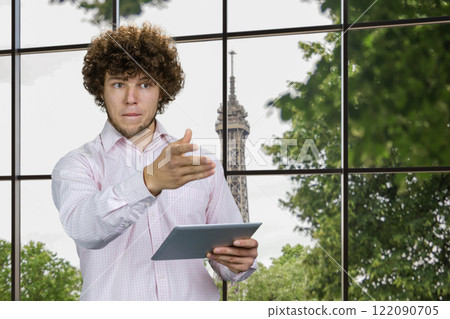 Portrait of a young caucasian worker with curly hair holding tablet pc device pointing at something. Checkered window with tv tower in the background. Portrait of a young caucasian worker with curly hair holding tablet pc device pointing at something. Checkered window with tv tower in the background. 122090705