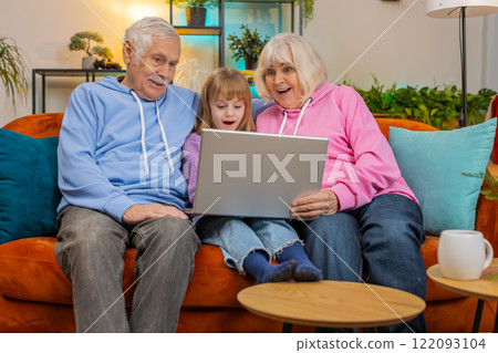 Senior couple man and woman with child girl granddaughter spending time at home sofa using laptop Senior couple man and woman with child girl granddaughter spending time at home sofa using laptop 122093104