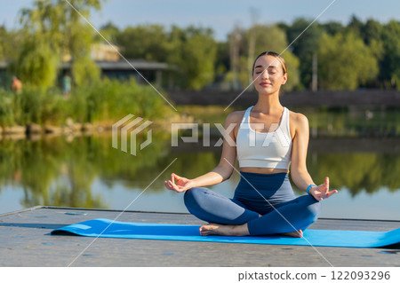 Caucasian young woman sitting on mat in lotus position, relaxing near pond in park on summer day Caucasian young woman sitting on mat in lotus position, relaxing near pond in park on summer day 122093296