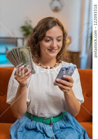Successful young woman holding smartphone and dollar bill money fan sitting on sofa couch at home Successful young woman holding smartphone and dollar bill money fan sitting on sofa couch at home 122093318