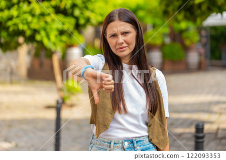 Upset young Caucasian woman lady showing double thumbs down expressing discontent on city street 122093353