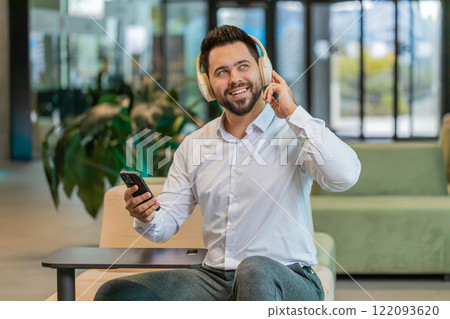 Excited young businessman in headphones listening energetic music on smartphone in office lobby Excited young businessman in headphones listening energetic music on smartphone in office lobby 122093620