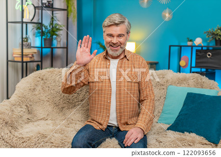 Mature man smiling friendly sitting on cozy sofa waving hand in greeting hello, hi at home room Mature man smiling friendly sitting on cozy sofa waving hand in greeting hello, hi at home room 122093656