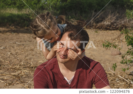 Young dad smiles as his young daughter, 4, covered his eyes with her palms on sunny day at beach in summer 122094876