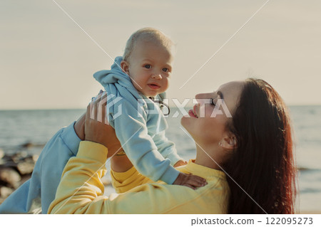 Baby in blue suit in arms of happy mum is lifted aloft against sea and sky on sunny day Baby in blue suit in arms of happy mum is lifted aloft against sea and sky on sunny day 122095273