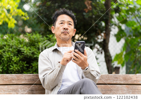 A middle-aged man sitting on a bench and operating a smartphone 122102438