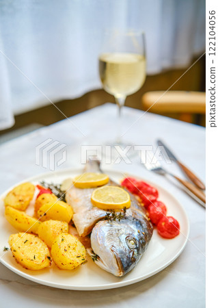 Freshly cooked dorado fish with lemon and roasted potatoes on a dining table with wine 122104056
