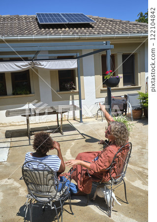 Two women sitting in a sunny backyard, pointing at solar panels Two women sitting in a sunny backyard, pointing at solar panels 122104842