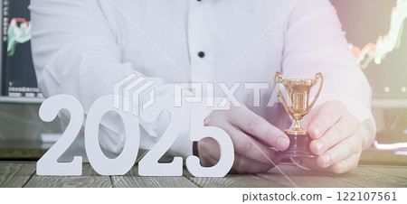 2025 business achievement and success concept: hands holding golden trophy with decorative numbers and stock charts on background 122107561