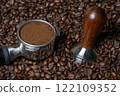 Coffee amper and a coffee portafilter filled with freshly ground coffee on a bed of many coffee beans 122109352