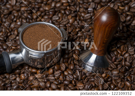 Coffee amper and a coffee portafilter filled with freshly ground coffee on a bed of many coffee beans 122109352