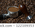 Coffee amper and a coffee portafilter filled with freshly ground coffee on a bed of many coffee beans 122109353