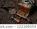 Rustic coffee grinder with a measuring spoon stands  with a tamper and a coffee portafilter filled with freshly ground coffee on a bed of many coffee beans 122109354