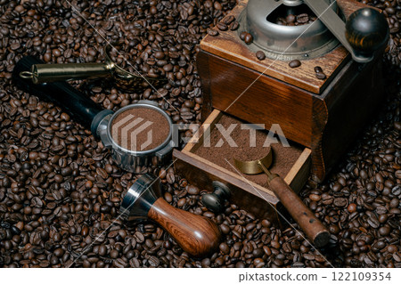 Rustic coffee grinder with a measuring spoon stands  with a tamper and a coffee portafilter filled with freshly ground coffee on a bed of many coffee beans 122109354