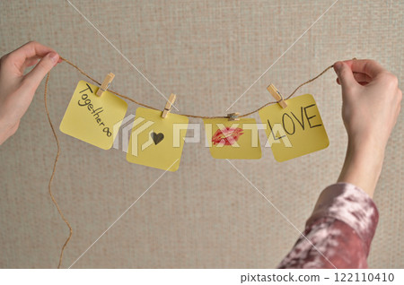 Woman's hands holding four yellow sticker papers with lipstick kiss mark, inscription "Love", "Together" and small black heart on a string with clothespins. Romantic message for Valentine's Day Woman's hands holding four yellow sticker papers with lipstick kiss mark, inscription "Love", "Together" and small black heart on a string with clothespins. Romantic message for Valentine's Day 122110410