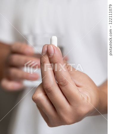 Woman in white t-shirt holding white pill in fingers and water glass in hand closeup 122111079