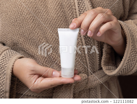 Closeup of woman in brown bathrobe holding squeeze cream tube in hands, cosmetic mockup Closeup of woman in brown bathrobe holding squeeze cream tube in hands, cosmetic mockup 122111082