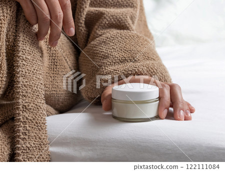 Woman in brown bathrobe puts white opened cream jar on white bedsheet closeup, mockup Woman in brown bathrobe puts white opened cream jar on white bedsheet closeup, mockup 122111084