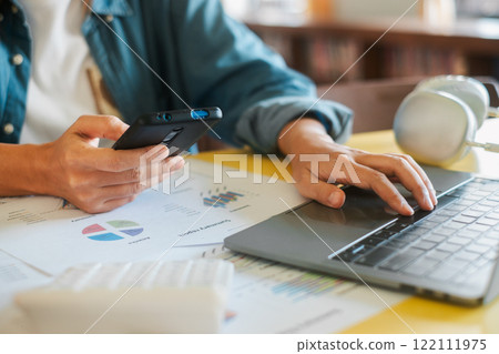 Close-up of a person using a laptop and smartphone to analyze financial charts and data in a modern office environment. Close-up of a person using a laptop and smartphone to analyze financial charts and data in a modern office environment. 122111975
