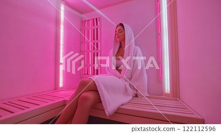 Young woman is sitting on a wooden bench, relaxing in an infrared sauna. 122112596