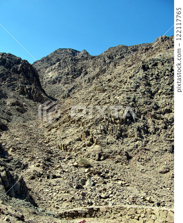 Narrow ascent to the mountain from a stone fence. Yellow and gray stones and blue sky 122117765