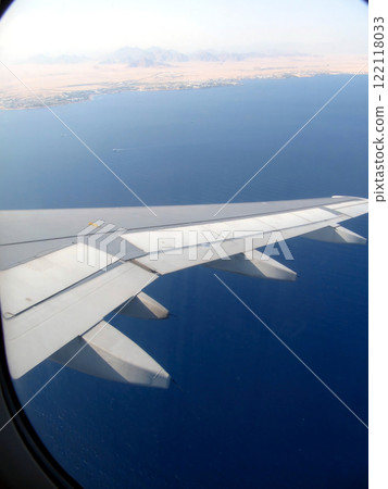 View From the Airplane Porthole Showcasing the Wing Over a Vast Blue Ocean and Land Nearby 122118033